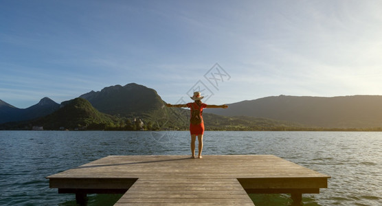 女孩站着戴一顶跟踪帽子穿着红色礼服双臂抬起看着山峰鞋子在码头上穿着湖边的生活方式概念春天日落船图片