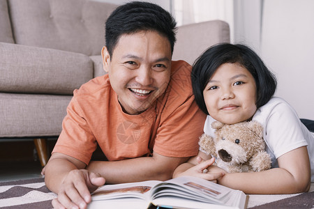 父母亚洲亲和女儿正在读书同时躺她家客厅的地毯上可爱的小女孩拥抱着娃与他父亲笑得开心快乐地微笑活的女士图片