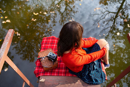 红色的一位年轻女子在阳光明媚的日子里背坐在湖边的一座桥上格子布相机手提箱一位年轻女子在阳光明媚的日子里背坐在湖边的一座桥上格子布图片