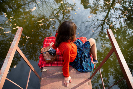 男人一位年轻女子在阳光明媚的日子里背坐在湖边的一座桥上格子布相机手提箱一位年轻女子在阳光明媚的日子里背坐在湖边的一座桥上格子布手图片