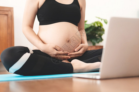 腹部在家做瑜伽的亚洲孕妇通过笔记本电脑在线视频培训课程在家里做瑜伽参加运动的女穿服触摸她们的肚子和在怀孕期间做锻炼健康的生活方式图片