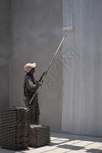 亚洲建筑工人的侧面视图使用长柄滚圈刷将白漆刻在垂直架子建房工地内水泥墙背景上的壁画底线在室内框架绘画图片
