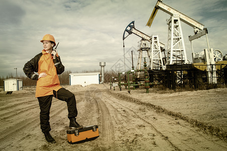 石油田的女工程师在电台上谈论时佩戴橙色头盔和工作服业场地背景油气工业人的背景情况TonedOilandgasIndustrywo图片