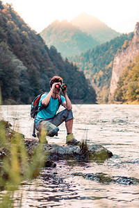 年轻的湿带着背包年轻旅游者在山峰上看望远镜站在河边的岩石上男孩在山度假与背包一起游荡他穿着运动夏季服装笑声保持图片