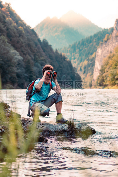 年轻的湿带着背包年轻旅游者在山峰上看望远镜站在河边的岩石上男孩在山度假与背包一起游荡他穿着运动夏季服装笑声保持图片