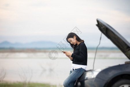 发动机男人坏了亚裔女在寻找时使用手机在街上因汽车故障而坐后图片