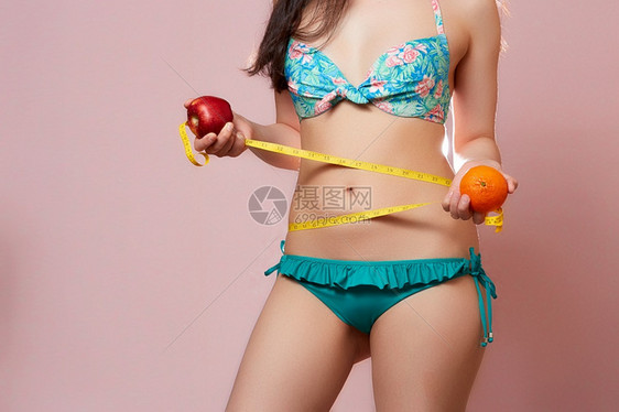 措施胖的身穿泳衣年轻苗头亚洲女孩一只手拿着橙子另一只手拿着苹果上测量胶带在粉红色孤立的背景上瘦身图片
