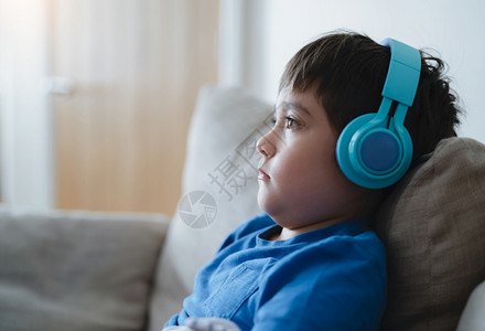 身穿耳机深思熟虑的男孩穿着蓝色T恤听音乐的学校男孩CuteKid坐在家中客厅的沙发上休息时唱歌在线的乐趣图片