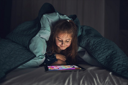 有趣的夜晚卧室女孩在看玩和听平板电脑上的故事时玩得开心孩子在睡前用平板躺床垫下之前玩得开心图片