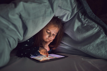 说谎童年就寝时间女孩在看玩和听平板电脑上的故事时玩得开心孩子在睡前用平板躺床垫下之前玩得开心图片