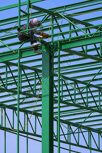 劳动者风镜亚洲建筑工人的低角度观点亚洲建筑工人在垂直框架蓝天空背景下将金属焊接在绿色屋顶仓库结构上的绿色屋顶壁架上风险图片