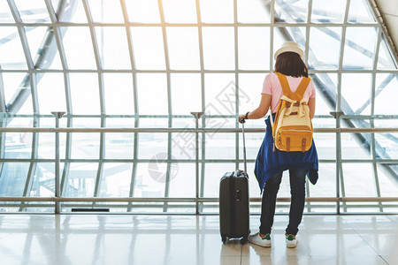 制作者帽子携带手提箱和黄色背包的亚洲年轻女子在机场后视镜窗口等待飞行的亚洲青年女子正在用智能手机空间检查飞行情况以观察飞行情况时图片
