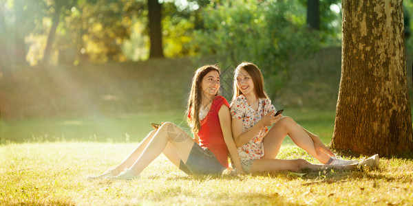 快乐的脸在明亮夏日两个美丽的青少年在一棵树下坐着Mobbil手机肖像图片