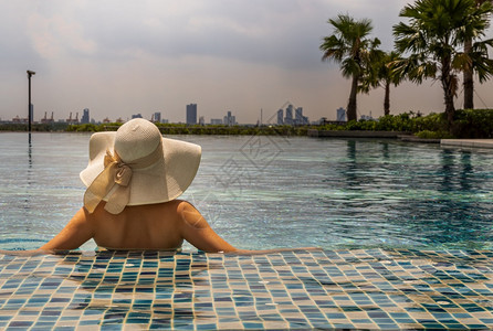 淑女在阳光明媚的日间暑假概念中年轻女在游泳池放松帽子时的回视日光浴城市的图片