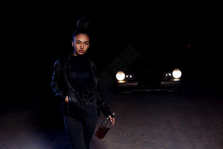 女一名身着皮夹克和黑衣服的非裔美国人穆拉托女孩正在一条废弃沙路上行走晚在汽车灯光和街道照亮下带着一瓶威士忌悲伤酒鬼图片