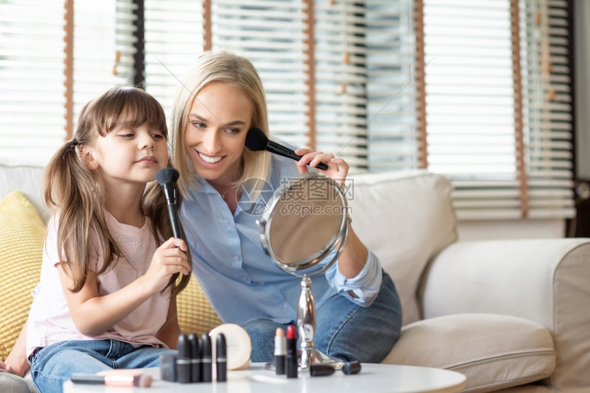 黑发微笑的母亲和女儿看着镜子在脸上涂刷子的化妆品年轻女人和小孩在家睡沙发时做善于安心快乐爱的家庭时候坐在沙发上裙子着图片
