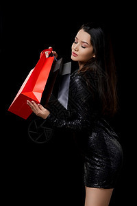 庆祝黑色的发一位身穿色连衣裙的年轻女子对礼物感到满意在黑色孤立的背景下买黑色和红的袋子图片
