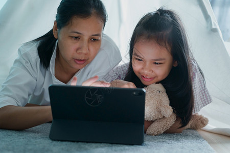 一种放松亚洲母亲和女儿的幸福家庭晚上在帐篷中使用平板电脑在亚洲孩子们拥抱一个洋娃聊天以及在家里数字电脑上学习技术图片