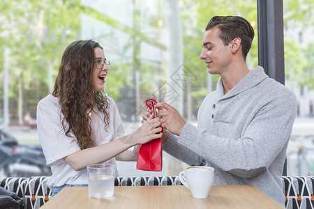 庆典约会吸引人的这是给你快乐男人给他爱妻送礼物女人满目欢喜地看着笑的情侣坐在餐厅桌前坐着图片