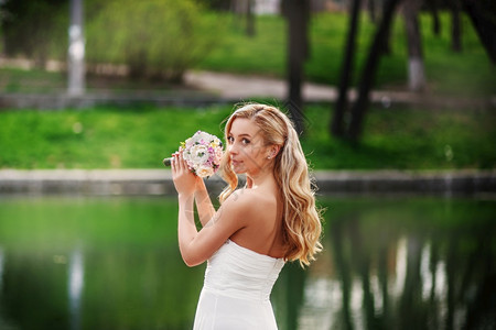 保持身着白裙子的年轻美丽新娘在水池附近站一束花上等待新郎漂亮的年轻图片