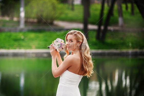 保持身着白裙子的年轻美丽新娘在水池附近站一束花上等待新郎漂亮的年轻图片