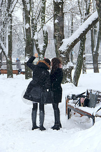 好的年轻女孩在白雪皑的冬季公园背景自拍及时冻结的女孩在冬季公园下雪时冬季公园现代女孩的自拍照朋友在冬天用智能手机自拍年轻女孩在白图片