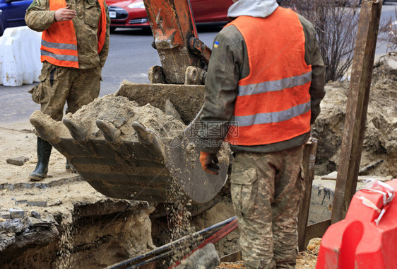 一桶被道路工人包围的重型建筑挖掘机在城市街道上进行下水维修时抬沙子被道路工人包围的重型建筑挖掘机桶在维修道路上的城市下水道时挖沟图片