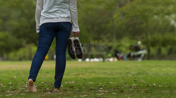 木头现代的绿色紧贴身穿蓝裤的年轻女子她们赤脚在公园的草地上走来去将运动鞋握在右手和绿树背景右手拿着她的运动鞋以及绿树背景在公园里图片