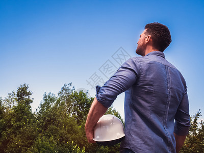 牛仔布保护的工程师英俊匠在绿树和日落太阳的背景下把白色硬帽放在公园里看着距离近的劳动和就业概念把白色硬帽子放在公园里b在绿色树木图片