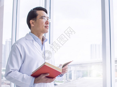 窗户男亚洲人身戴眼镜学习和阅读书籍的亚洲聪明医生站在窗口教育和职业概念旁边身着眼镜学习和阅读书籍的亚洲智能医生图片