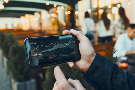 偏僻的使用智能手机上的投资应用程序在股票加密货币市场上投资交易的人手持机交易者查看蜡烛图使用智能手机上的投资应用程序在股票加密货背景图片
