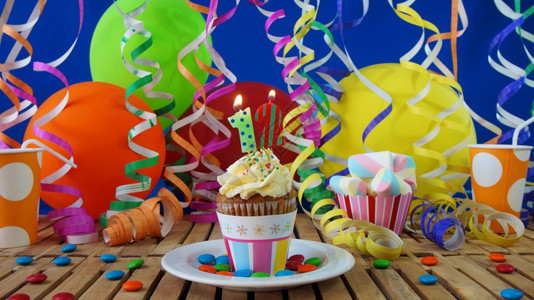节日生12小蛋糕在质朴的木桌上燃烧着蜡烛背景是五颜六色的气球塑料杯和糖果背景是蓝色的派对墙生日的蛋糕背景是五颜六色的气球塑料杯和图片