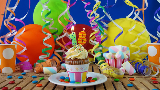 天木制的幸福18岁生日小蛋糕在质朴的木桌上燃烧着蜡烛背景是五颜六色的气球塑料杯和糖果背景是蓝色的派对墙背景图片