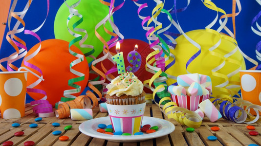 庆典杯子祝活动19岁生日小蛋糕在质朴的木桌上燃烧着蜡烛背景是五颜六色的气球塑料杯和糖果背景是蓝色的派对墙背景图片