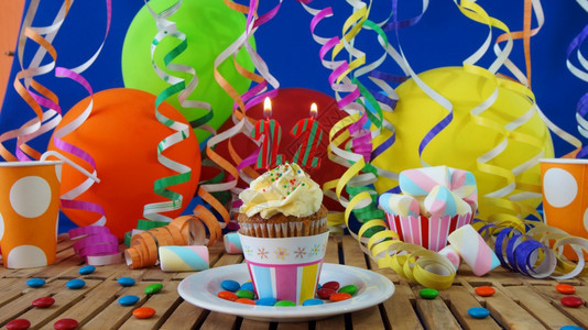 生日2小纸杯蛋糕在质朴的木桌上燃烧着蜡烛背景是彩色气球塑料杯和糖果背景是蓝色派对墙纸杯蛋糕背景是五颜六色的气球塑料杯和糖果背景墙图片