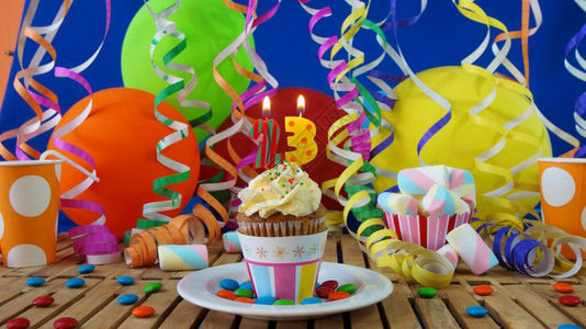 惊喜杯子生日23小纸杯蛋糕在质朴的木桌上燃烧着蜡烛背景是五颜六色的气球塑料杯和糖果背景是蓝色的派对墙背景展示图片