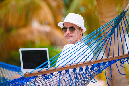 年轻人在海滩度假的吊床上放松与膝型笔记本电脑一起在热带度假的吊床上笔记本电脑的青年男子户外棕榈游客图片