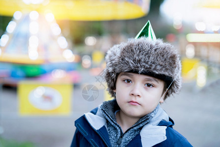 积极的家庭可爱孩子带着毛的党帽看着深沉的思考年轻男孩独自站在外面边与模糊的彩色灯光在游乐场背景中模糊的彩色灯光孩子在冬季节日学校图片