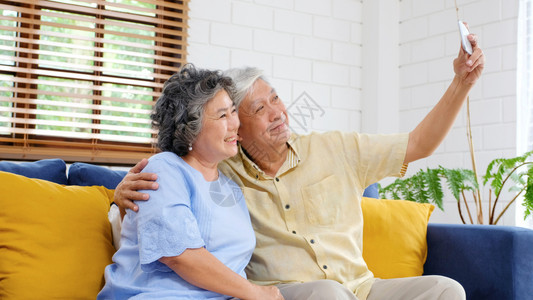 快乐的老年亚裔夫妇在家里客厅自拍在欢乐时刻活跃的老年人有技术和生活方式的临时退休者以及具有技术和生活方式的临时退休者手机女愉快图片