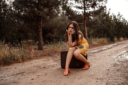 成人常设身着黄色裙子的年轻美女穿着彩色围巾坐在雨后湿泥路上的旧式手提箱女士图片