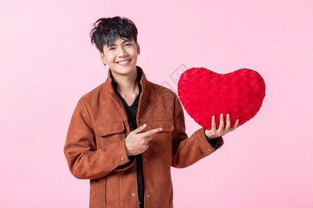 快乐的亚洲男子英俊年轻拿着红色心形枕头在爱情中被孤立粉红空白间室背景的照本上CED保持礼物图片