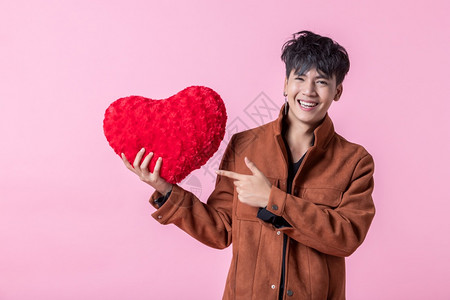 亚洲男子英俊年轻拿着红色心形枕头在爱情中被孤立粉红空白间室背景的照本上CED愉快情人节假期图片