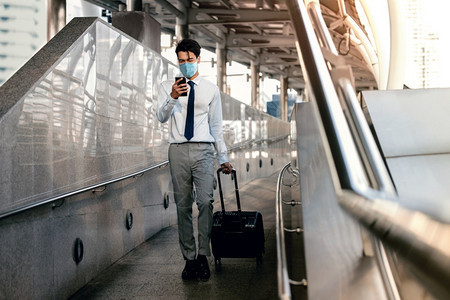 年轻亚洲商人在机场或公共交通站健康护理新正常生活方式概念中穿着外皮面罩和使用智能电话与手提箱一起行走时使用智能电话的新型普通生活图片