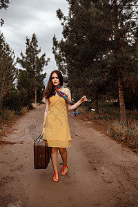 奢华成人身着黄裙子的年轻美女围巾颜色多彩穿着旧式手提箱在雨后湿泥地路上头发图片