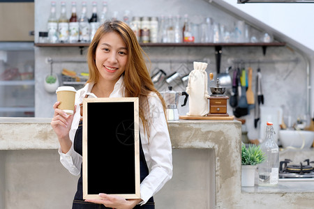 展示年轻的女亚洲咖啡师拿着一次咖啡杯和空黑纸板在咖啡馆的柜台背景食物和饮料概念上面带微笑的脸曼谷店铺图片