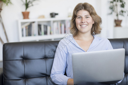 技术打字随意的身着蓝衬衫企业家妇女在沙发上用笔记本电脑工作图片
