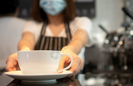 围裙身戴面罩的亚洲女服务员妇为预防传染病而佩戴面罩并举着咖啡杯为店顾客提供服务19世纪COVID爆发后的安全和预防概念杯子保持图片