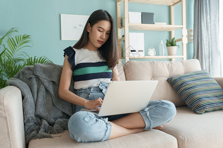 人们在家中客厅沙发上坐着电脑膝型笔记本电脑的女青年早上做打字工作的Asian女孩她在家里工作活的女图片