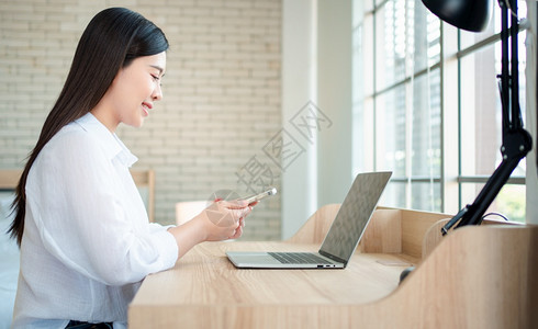 年轻的幸福快乐美丽亚洲女在家庭办公室用笔记本电脑和智能手机工作坐在桌子上的主机办公室使用笔记本电脑和智能手机概念上网的女职业自由图片
