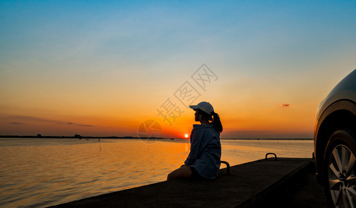 美丽的孤单年轻女子休丽月光在沙滩上戴着帽子独自在车前放松日落暑假和旅行概念下黄橙蓝天空随意的坐图片